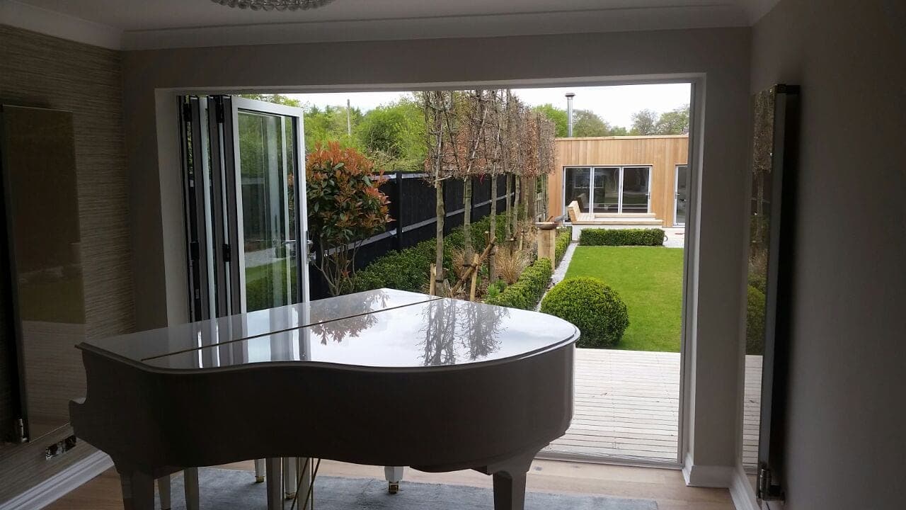 Aluminium bifold doors from Cortizo opening onto a garden with a grand piano in view